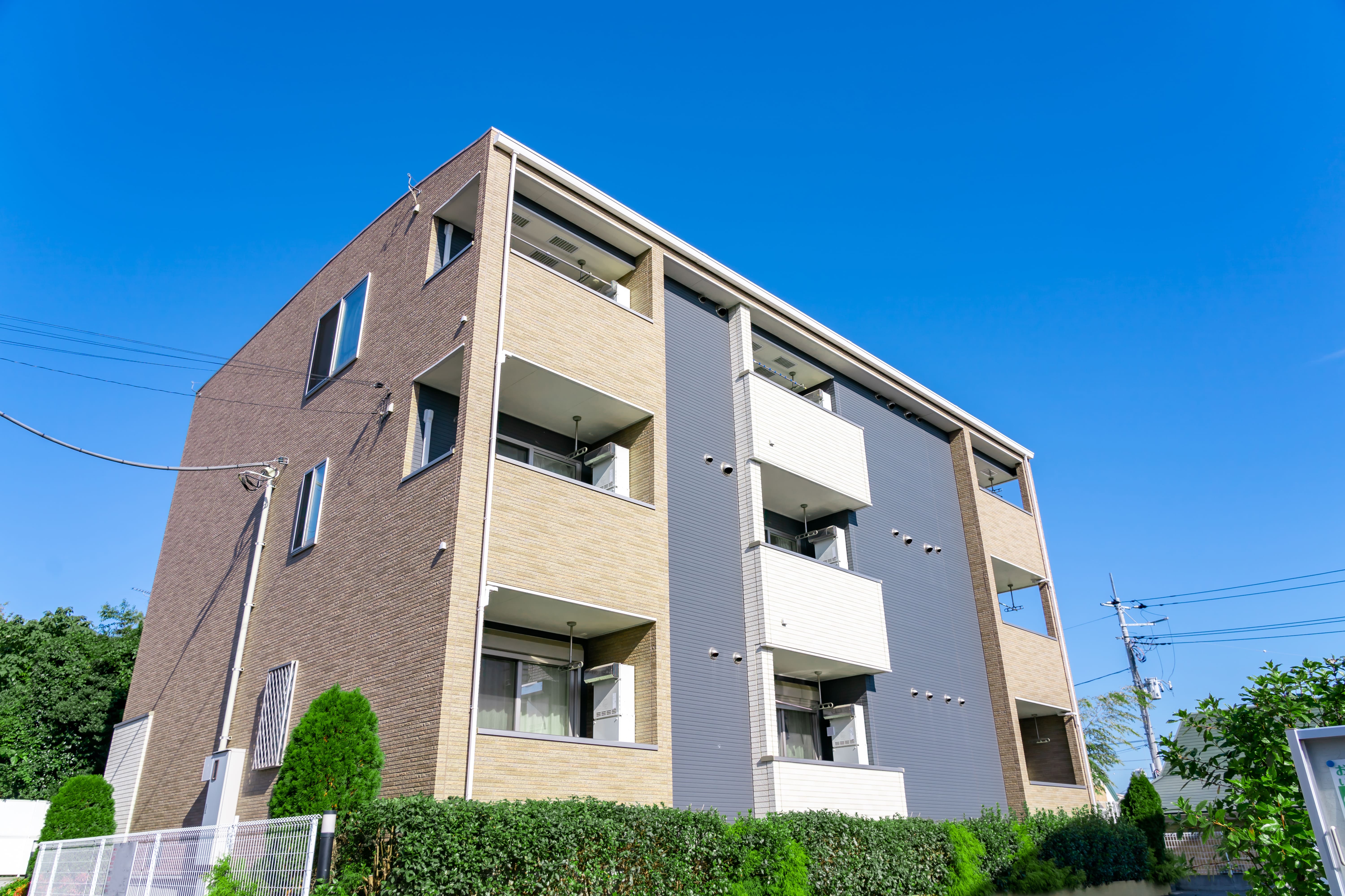 青空の下、茶色とグレーの外壁のアパートメントと植え込み。